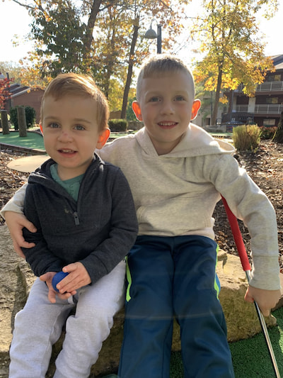 Two young boys, one with arm around the other, sit together on a miniature golf course