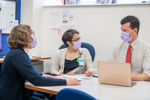 Two women and one man, all wearing surgical masks, sit together at a conference table.