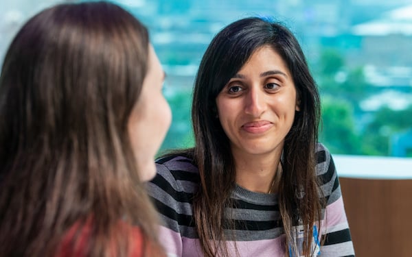 A woman facing the camera is in conversation with another woman with a bright window in the background.