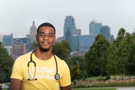 Ijon Howard smiling, and standing outside with the Kansas City, Mo. skyline behind him. He is wearing a stethoscope around his neck and a shirt that reads, " I LOVE tiny humans."