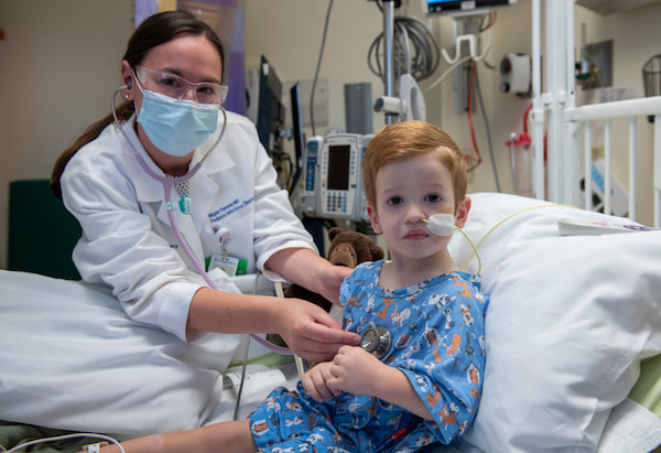 A female physician uses a stethoscope on a young male child who has a feeding tube.