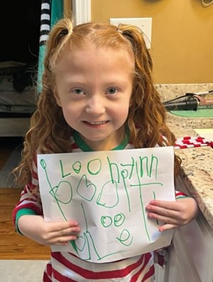 A young girl with red hair in pigtails holds up a piece of artwork she has created.