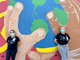 Abby Kietzman and Shannon Hill, RN, BSN wearing facemasks and standing outside, in front of a mural with three hands holding the planet Earth.