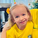 A young blond girl in a yellow t-shirt with a yellow bow in her hair smiles for the camera