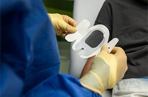 Two gloved hands placing a clear sticker on a young patient's arm to keep a PICC line clean and safe.