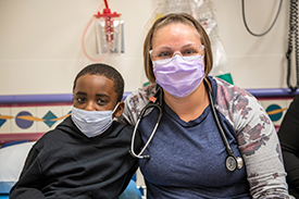 Chase Jackman with nurse, Lindsey Fricke. They have their arms around each other and are wearing face masks inside a Children's Mercy patient room.n's Mercy patient room.