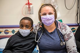 Chase Jackman with nurse, Lindsey Fricke. They have their arms around each other and are wearing face masks inside a Children's Mercy patient room.n's Mercy patient room.