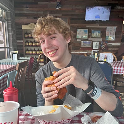 A teenage boy sits at a restaurant table holding a chicken sandwich, with a food basket and drink on the table.