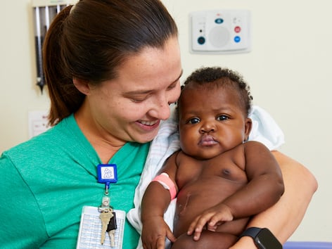A female health care worker in a green shirt gently holds a Black baby girl during a clinical visit, with medical equipment visible in the background.