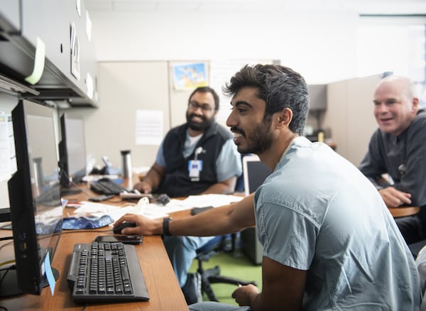 A young man in scrubs looks at a computer monitor as two other men look on.