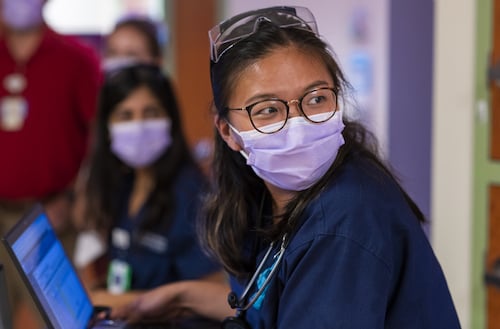 A young woman wearing a surgical mask, glasses, goggles on her head and scrubs looks over her shoulder while working with a laptop computer.