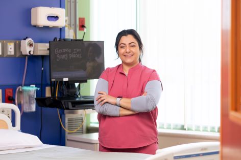 A woman wearing pink scrubs is smiling with her arms crossed in a hospital room.