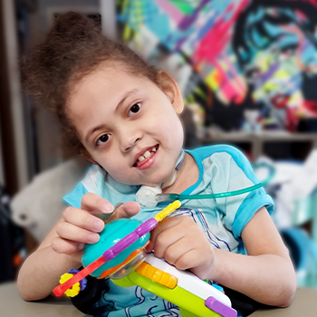 Children's Mercy patient, Celia. She has a trach tube, is smiling and playing with a toy.