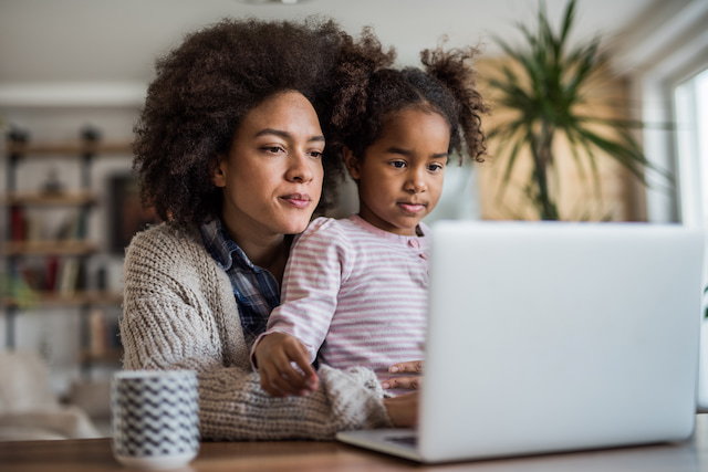 A Black woman with a young girl on her lap looks at a laptop computer.