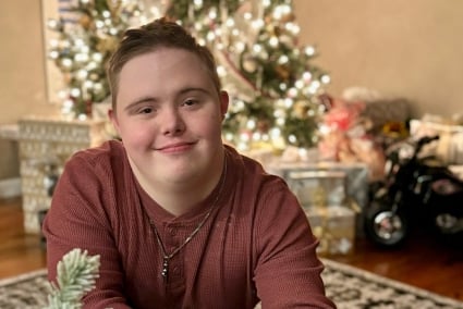 A young man with brown hair in a burgundy shirt sits on the floor in front of a Christmas tree.