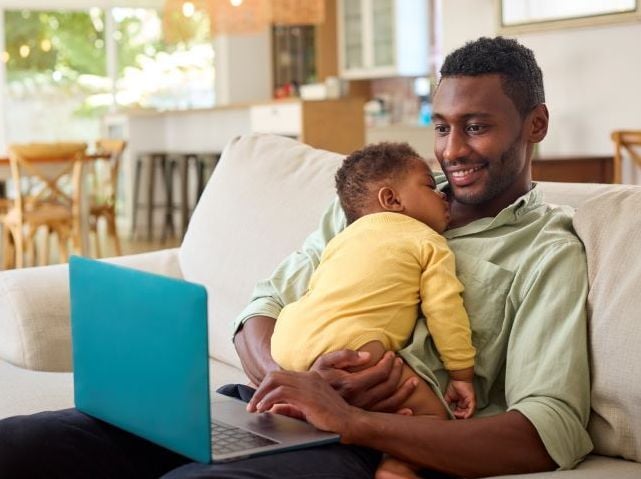 A Black father sits on a couch holding a sleeping baby while using a laptop at home.