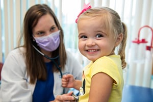 Blond-haired female toddler in yellow shirt smiles at camera while being examined by a Children's Mercy resident.
