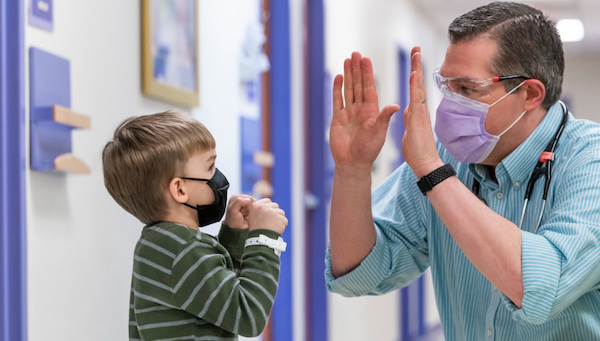 A young boy wearing a face mask high fives a masked adult man with a stethoscope around his neck.