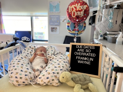 A preemie baby is propped on a pillow in a NICU crib with a sign that reads “Due dates are so overrated! Franklin Wayne.”