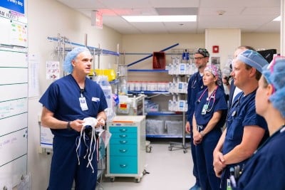 A male physician in blue scrubs speaks to a group of male and female physicians in a hospital setting.