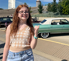  Lacie outside at Gateway Arch National Park. She is smiling and pointing over her shoulder at a green classic car.