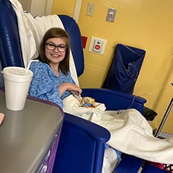 Taylor Stewart smiling while sitting in a blue hospital chair wearing eyeglasses and a blue hospital gown. She has a white blanket over her legs and has Goldfish crackers in her lap.