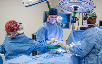 Two surgery fellows assisting in a surgery with a Children's Mercy doctor. One fellow is handing the doctor a medical instrument. All are wearing surgical protective items, including face masks, surgery caps, gowns, and gloves.