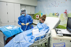 A A Children's Mercy nurse wearing a blue gown, gloves, and a hat  and a young patient wearing a face mask and covered with a blue sheet.