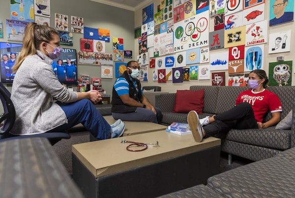 Three masked young adults chat in a hospital break room.