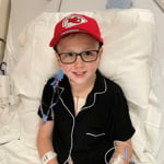 A young boy in glasses and a Chiefs hat sits in a hospital bed