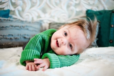 A baby boy with spiky hair and a green sweater crawls on his stomach.