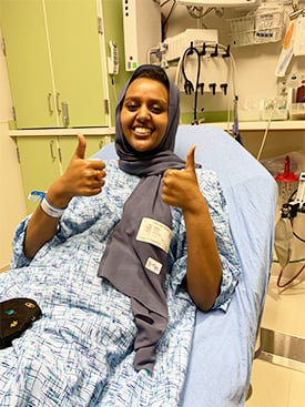 Munira Nuru on a gurney at Children's' Hospital, smiling and give two thumbs-up.