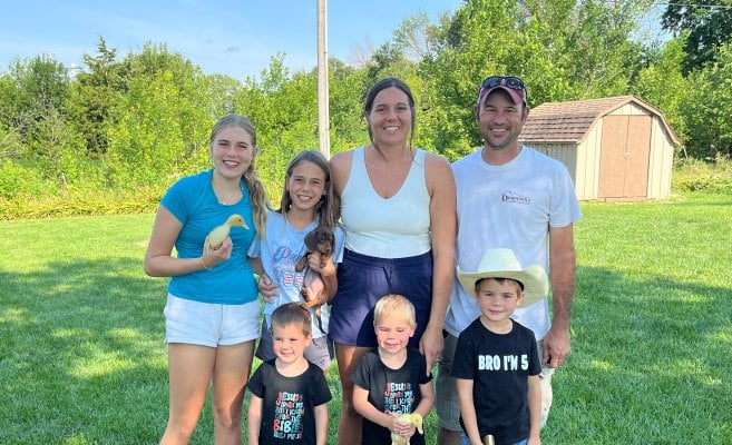 A family poses for a photo in a large green yard. There are trees and a small shed behind them. In the back row are two older girls with a mom and dad. One girl is holding a yellow duckling and the other is holding a puppy. In the front row are two toddler boys (one is holding a duckling, too) and a little boy in a cowboy hat holding a toy gun. He’s wearing a t-shirt that reads “Bro I’m 5.” 
