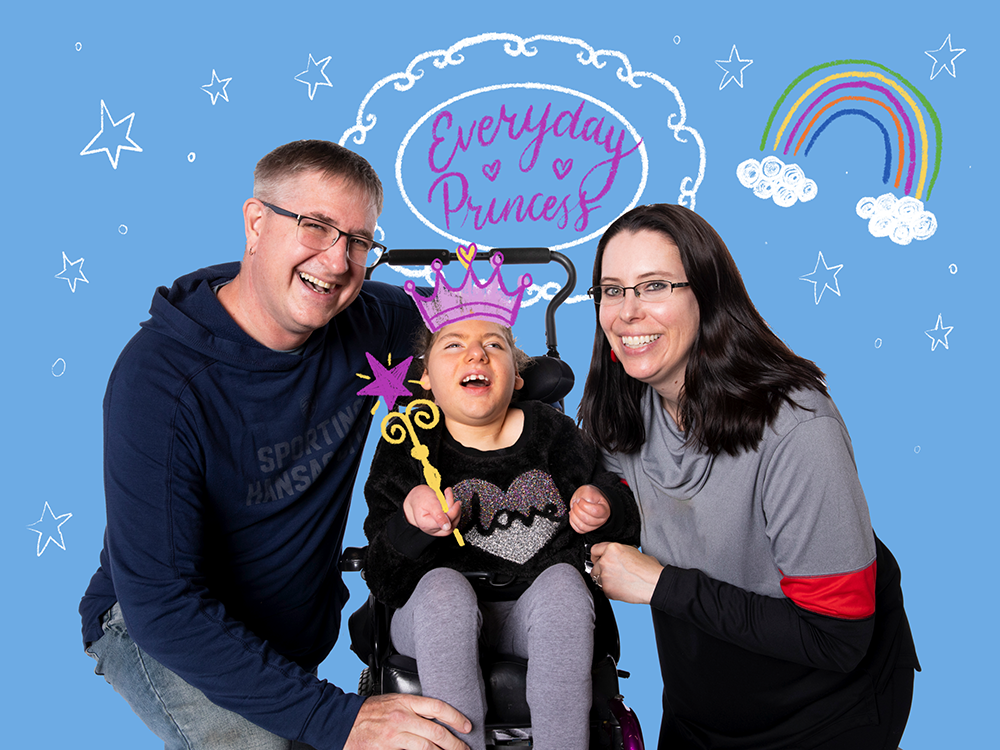 Hudsyn, a patient with complex medical needs, with her parents. She's holding an illustrated wand and she's wearing an illustrated crown. Light blue background with stars, a rainbow, and "Everyday princess."