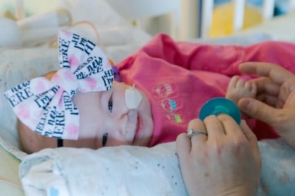 A smiling baby girl with a large bow on her head and a tube leading to her nose lays on her side and smiles. An adult holds her hand and a blue pacifier.