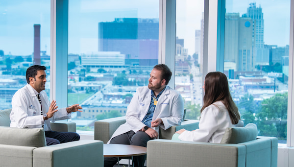 Two men and one woman, all in white coats, sit talking together near a window overlooking Kansas City.