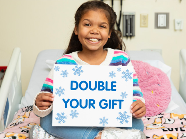 Children's Mercy patient, Tristyn, is sitting on a Children's Mercy hospital bed, smiling, and holding a sign that read, "double your gift." 