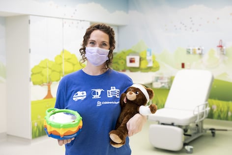 Lenore Fleming,a Children's Mercy Child Life Specialist, wearing a mask and holding a teddy bear and a child's drum.