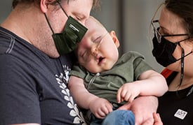 Odin Buser wearing a nasal cannula and being cradled by his father and mother who are both wearing face masks.