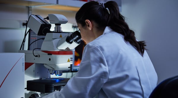 A dark-haired woman with a ponytail and a lab coat looks into a microsope.