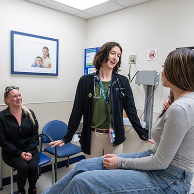 A tall, female doctor with chin-length brown hair stands facing a patient. The patient is a teenage girl with long dark hair with blonde highlights. She is wearing a gray shirt and jeans and sitting on an exam table. The patient’s mom sits off to the side, smiling and watching the interaction.