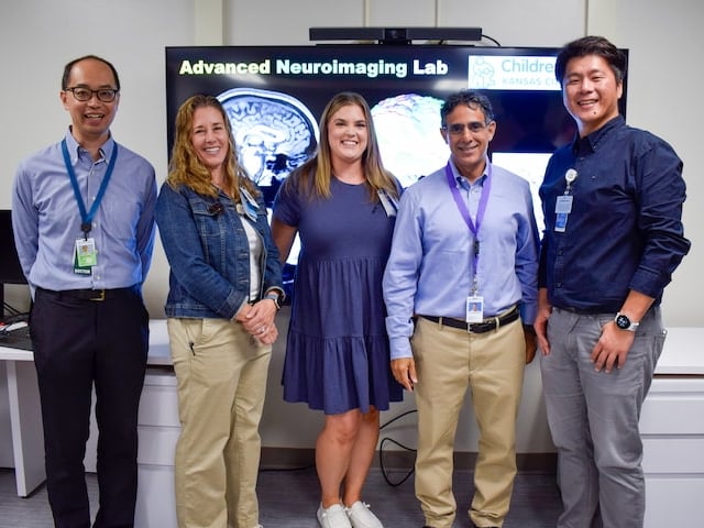 Five staff members stand together in a laboratory in front of a large display showing brain scan images. The screen behind them reads “Advanced Neuroimaging Lab” , and the group is posing in a bright clinical research space.