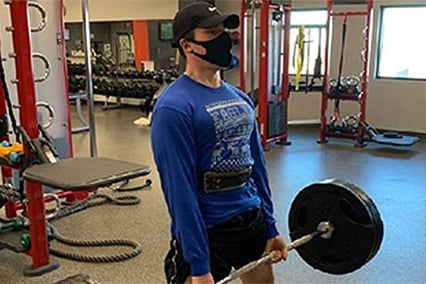 Gabe Keeton wearing a black face mask and lifting a barbell in a gym. 