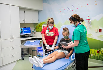 Girl sitting on a hospital bed while a child life specialist relaxes her with an iPad and a nurse cleans the girl's burn.