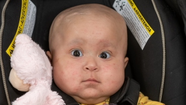 A baby with chubby cheeks and a pink stuffed animal looks directly at the camera.
