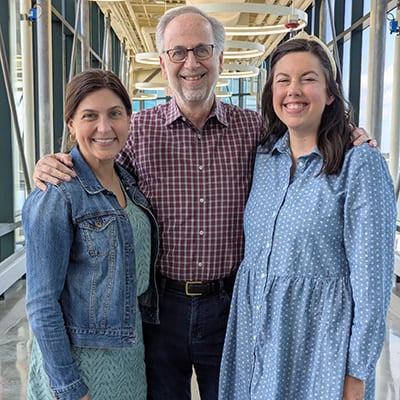Three adults stand together in a hospital corridor with windows and overhead lighting.