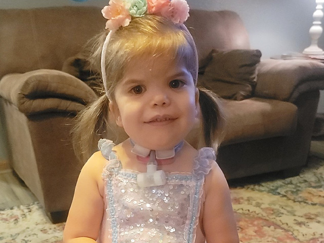  Emersyn Gross at home smiling with her hair in pig tails, wearing a sparkly dress and flower headband.