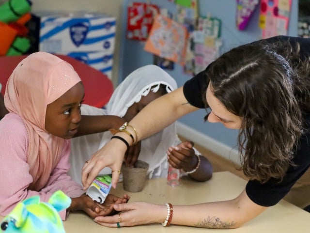 An adult helps two children with a hands-on activity at a classroom table.