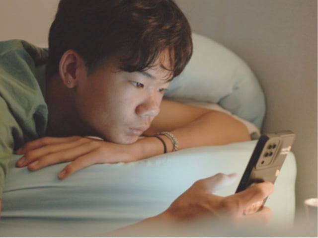 An Asian teenage boy lies on a bed while scrolling through a cell phone.