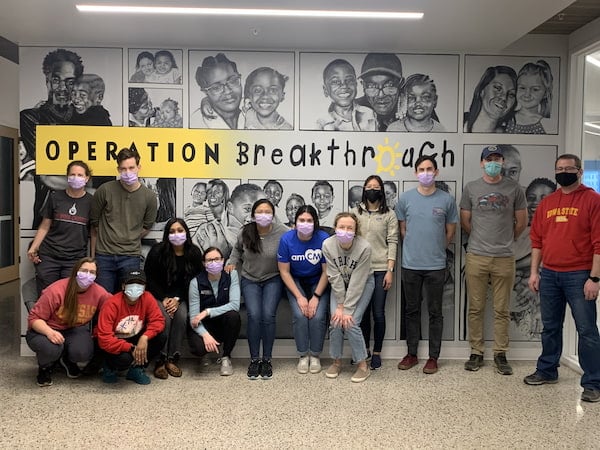 Several masked young adults pose together near an Operation Breakthrough sign
